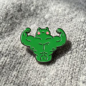 May include: A green enamel pin shaped like a muscular frog with defined abdominal and arm muscles. The frog has a small head, a flat mouth line, and two round pink spots on its cheeks. The design features black outlines and a glossy finish.