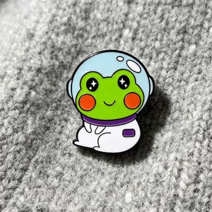 Peut inclure: Une broche en émail métallique représentant une grenouille vert vif portant une combinaison d'astronaute blanche avec une visière de casque bleu clair. La grenouille a de grands yeux noirs avec des reflets d'étoiles, des joues orange rondes.