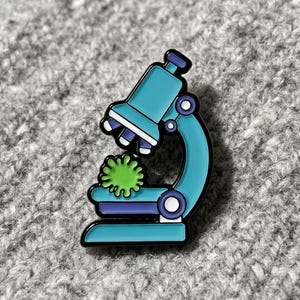 Könnte beinhalten: Ein Emaille-Pin mit einem türkis-violetten Mikroskop und einem grünen Virus-Design. Das Mikroskop hat weiße Akzente und eine schwarze Umrandung. Der Pin befindet sich vor einem grau strukturierten Hintergrund.