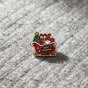 May include: A red enamel pin shaped like a sleigh carrying a green Christmas tree, wrapped gifts, and a gnome wearing a red hat. The sleigh features a black sign with the white text Merry Christmas and gold trim accents against a red background.