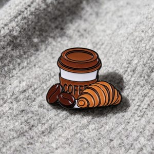 May include: A metal enamel pin featuring a brown and white coffee cup with the word COFFEE printed on the sleeve, accompanied by two dark brown coffee beans and a golden-brown croissant, all outlined in black metal against a grey background.
