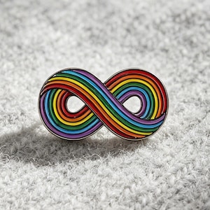 May include: A colorful enamel pin in the shape of an infinity symbol. The pin features a rainbow design with red, orange, yellow, green, blue, and purple stripes. The pin is set against a gray, textured background.