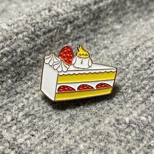 Peut inclure: Une broche en émail métallique en forme de part de gâteau aux fraises avec des couches jaunes, un glaçage blanc et un petit oiseau calopsitte jaune et blanc perché sur le dessus. La part de gâteau présente des fraises rouges et de la crème.