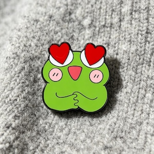 Könnte beinhalten: Eine grüne Frosch-Emaille-Anstecknadel mit zwei großen roten herzförmigen Augen, einer kleinen rosa dreieckigen Nase, zwei hellrosa runden Wangen und zwei vor der Brust gefalteten Händen, alles umrandet von einem kräftigen schwarzen Metallrand.