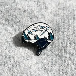 Puede incluir: Un pin de esmalte metálico con forma de casco de esquí sobre fondo blanco que presenta una ilustración gráfica de montañas nevadas, pinos de color verde azulado oscuro y una sección inferior azul oscuro con detalle de hebilla y contornos negros.