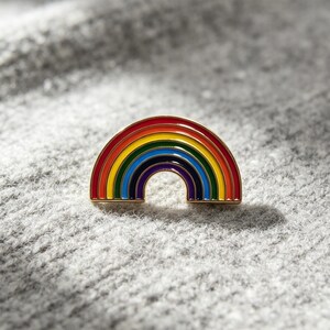 Puede incluir: Un broche de esmalte y metal dorado con forma de arcoíris semicircular. El diseño presenta bandas concéntricas de esmalte rojo, naranja, amarillo, verde, azul y morado, separadas por finas líneas de metal dorado, creando un aspecto pulido y brillante.