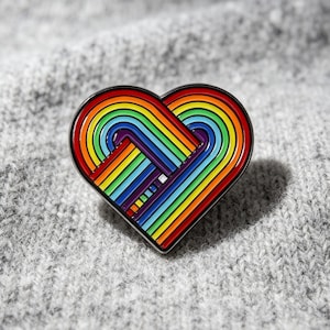 May include: A heart-shaped enamel pin with a rainbow design. The pin features a black outline and a vibrant array of colors, including red, orange, yellow, green, blue, and purple. The pin is set against a gray, textured background.