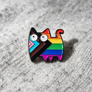 Peut inclure: Une broche en émail métallique en forme de chat stylisé. Le chat présente un design coloré avec des rayures horizontales rouges, orange, jaunes, vertes, bleues et violettes, ainsi qu'une section triangulaire bleu clair, rose, blanche, brune et noire.