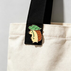 May include: A cute enamel pin of a hedgehog with a green leaf hat. The hedgehog has a tan body and brown quills. The pin is attached to a black tote bag strap. The tote bag is cream-colored.