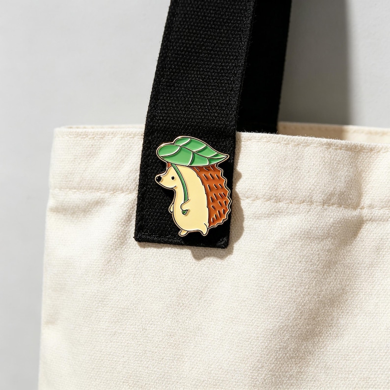 May include: A cute enamel pin of a hedgehog with a green leaf hat. The hedgehog has a tan body and brown quills. The pin is attached to a black tote bag strap. The tote bag is cream-colored.