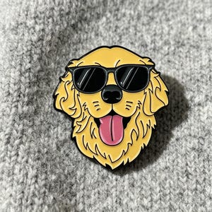 May include: A yellow enamel pin featuring a Golden Retriever dog wearing black sunglasses. The dog has a pink tongue sticking out. The pin is set against a gray, textured background.
