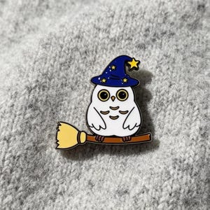 Könnte beinhalten: Eine Emaille-Anstecknadel in Form einer weißen Eule mit einem dunkelblauen Zaubererhut, der mit gelben Sternen und einer gelben Mondsichel verziert ist. Die Eule sitzt auf einem braunen Besenstiel mit gelbem Strohende im Cartoon-Stil.
