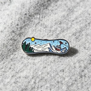 May include: An enamel pin featuring a horizontal landscape scene with snow-capped mountains, dark green pine trees, a bright yellow sun, and a red cable car suspended against a light blue sky with small white snowflakes scattered throughout.