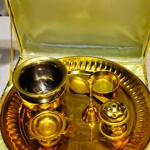 May include: A gold-colored metal puja thali set presented in a yellow satin-lined box. The set includes a round tray with embossed script, a small bowl, a diya lamp, a bell with a handle, and a small incense holder, all in a polished gold finish.