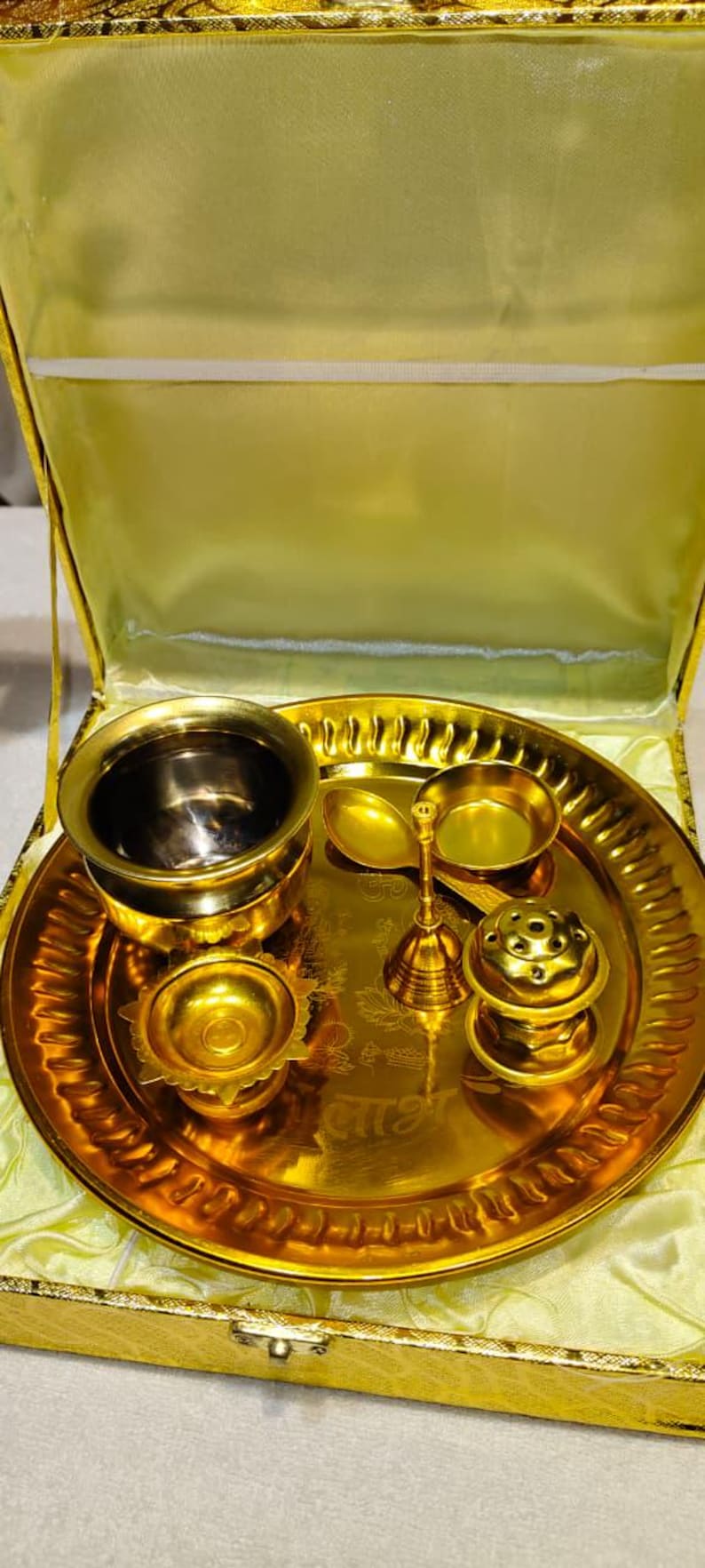 May include: A gold-colored metal puja thali set presented in a yellow satin-lined box. The set includes a round tray with embossed script, a small bowl, a diya lamp, a bell with a handle, and a small incense holder, all in a polished gold finish.