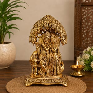 May include: A gold-toned metallic figurine depicting two figures standing together under a canopy of leaves. One figure holds a flute, while a cow stands beside them. The sculpture rests on a rectangular base with intricate decorative carvings.
