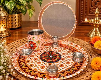 Puede incluir: Una bandeja redonda de metal con un patrón floral blanco y naranja y un diseño de mandala central, acompañada de una taza de metal a juego, un recipiente pequeño con tapa, dos cuencos plateados y un tamiz circular con borde rosa.