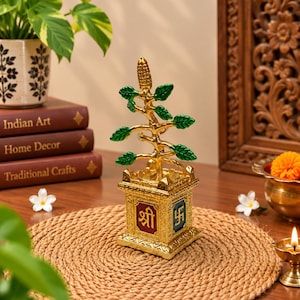 May include: A gold-toned metal Tulsi plant sculpture featuring green leaves and a central corn-like element. The plant sits in a square base with red and blue panels displaying the Devanagari script 'Shri' and the Swastika symbol in gold relief.