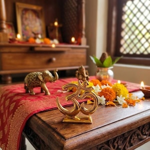 Könnte beinhalten: Ein goldenes Om-Symbol auf einem Holztisch, umgeben von Ringelblumen und Kerzen. Ein kleiner Messingelefant und eine Gottheitsstatue sind ebenfalls vorhanden, mit einem roten Stoff mit Muster darunter.