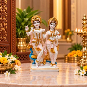 May include: A white marble-finish statue of Radha and Krishna standing together on a rectangular base. The figures are adorned with gold-toned jewellery, crowns, and draped garments with gold accents. A small blue peacock stands beside Krishna.
