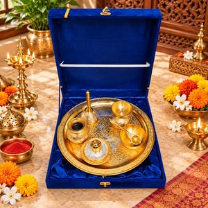 May include: A gold-toned metal puja thali set presented in a deep blue velvet-lined box. The set includes a circular tray with intricate engravings, a small bell, a diya, a bowl, and a spoon, all featuring matching metallic gold finishes.