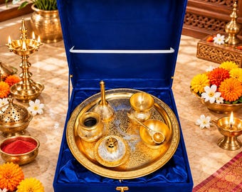 Puede incluir: Un juego de puja thali de metal dorado presentado en una caja forrada de terciopelo azul intenso. El conjunto incluye una bandeja circular con grabados intrincados, una pequeña campana, una diya, un cuenco y una cuchara dorados.