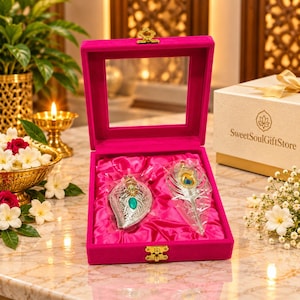 Puede incluir: Una caja de regalo de terciopelo rosa brillante con tapa transparente que contiene dos objetos decorativos plateados con forma de plumas de pavo real. Uno tiene una piedra verde y el otro un detalle de ojo de pluma.