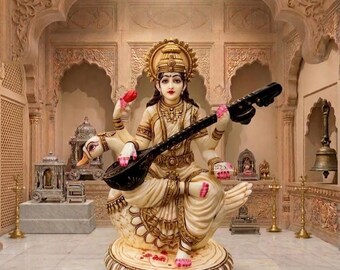 May include: A cream-colored statue of the Hindu goddess Saraswati, depicted with four arms, seated on a swan, and holding a black veena. The figure wears ornate gold-toned jewelry and a draped garment, positioned on a circular lotus-shaped base.