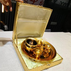 May include: A gold-colored metal puja thali set presented in a matching gold-toned hinged box with a satin-lined interior. The set includes a circular tray, a small bowl, a bell, and other ritual vessels, all featuring a polished, reflective finish.