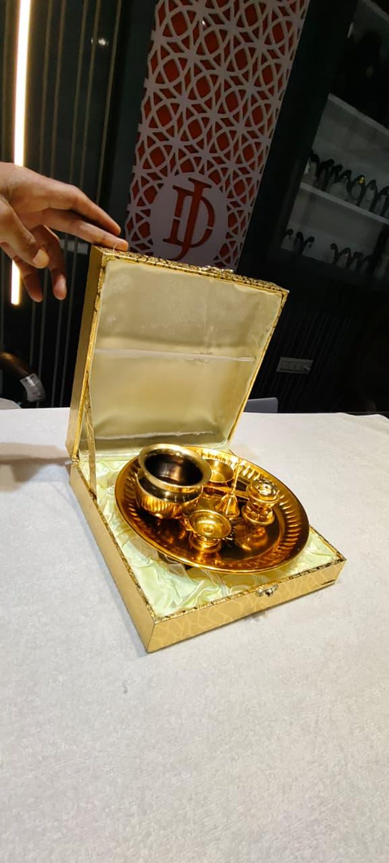May include: A gold-colored metal puja thali set presented in a matching gold-toned hinged box with a satin-lined interior. The set includes a circular tray, a small bowl, a bell, and other ritual vessels, all featuring a polished, reflective finish.