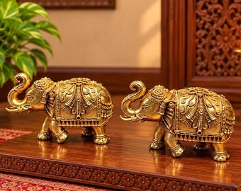 Puede incluir: Dos figuras de elefantes de color dorado con intrincados patrones en relieve en sus cuerpos y mantas. Cada elefante está representado con la trompa levantada, sobre una base rectangular de madera con detalles florales tallados.