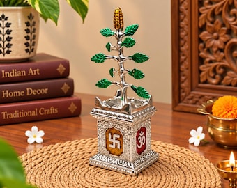 Puede incluir: Una escultura de planta Tulsi de metal plateado con hojas verdes y un capullo dorado en la parte superior. La base cuadrada está decorada con patrones intrincados y dos paneles insertados, uno con una esvástica amarilla y el otro con un símbolo Om rojo.