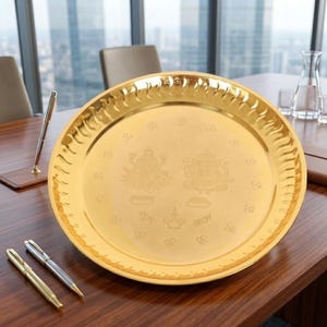 May include: A round, polished gold-colored metal tray featuring an embossed design in the center. The engraving depicts two deities, a diya lamp, and script text. The tray has a wide, scalloped rim with a repeating wave-like pattern along the edge.