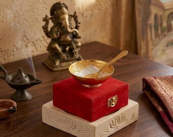 May include: A brass Ganesha statue, a small brass oil lamp, and a red velvet box with a gold-tone metal latch. A gold-tone bowl with a matching spoon rests on the box, which sits atop a square, light-colored stone base with carved lotus motifs.