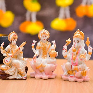 May include: Three small, white and gold statues of Hindu deities. The statue on the left depicts a figure playing a stringed instrument. The center statue shows a figure holding a pot. The statue on the right depicts a figure with an elephant head.