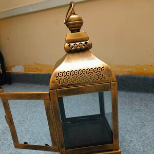 Puede incluir: Un farol de metal dorado con acabado envejecido, que cuenta con una puerta abatible abierta. El farol tiene una base cuadrada, paneles de vidrio transparente y una secci&oacute;n superior decorativa con un asa de anilla y patrones geom&eacute;tricos.