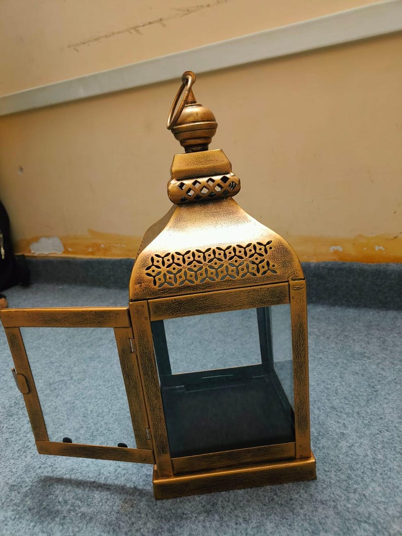 Puede incluir: Un farol de metal dorado con acabado envejecido, que cuenta con una puerta abatible abierta. El farol tiene una base cuadrada, paneles de vidrio transparente y una secci&oacute;n superior decorativa con un asa de anilla y patrones geom&eacute;tricos.