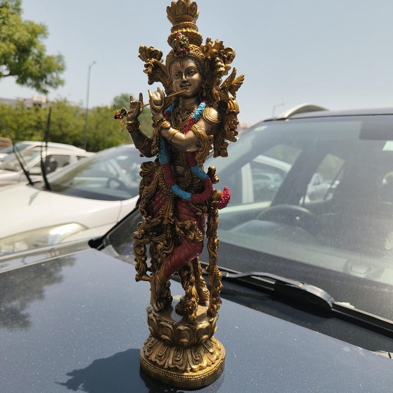 Puede incluir: Estatua detallada de una deidad, con adornos dorados y bronce. La figura est&aacute; adornada con una corona, joyas y una prenda roja y azul fluida. La estatua est&aacute; posando tocando una flauta, de pie sobre una base decorativa.