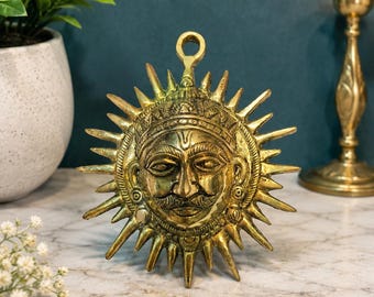 Puede incluir: Un adorno de pared circular de latón que presenta un rostro humano central con bigote, rodeado de rayos de sol radiantes. La pieza tiene una pequeña anilla en la parte superior para colgarla y muestra detalles grabados en el rostro.