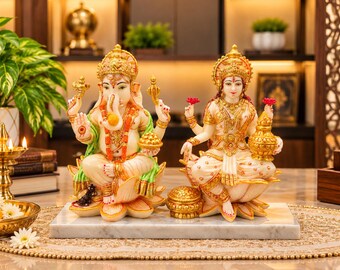 Puede incluir: Un par de estatuas blancas y doradas que representan a Ganesha y Lakshmi, ambas sentadas sobre bases de flor de loto. Ganesha tiene cabeza de elefante y cuatro brazos, mientras que Lakshmi sostiene una vasija dorada sobre una base de mármol.