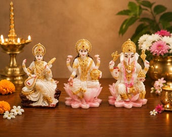Puede incluir: Tres estatuas blancas y doradas de deidades, incluyendo a Saraswati tocando una veena, Lakshmi sosteniendo una vasija y Ganesha, cada una adornada con intrincadas joyas y coronas doradas, sobre una superficie de madera con flores y una lámpara.