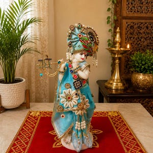 May include: A decorative figurine of a deity dressed in a light blue garment with gold trim, white shell accents, and colorful jewels. The figure wears an ornate blue turban and holds a gold flute adorned with small blue and green stones.