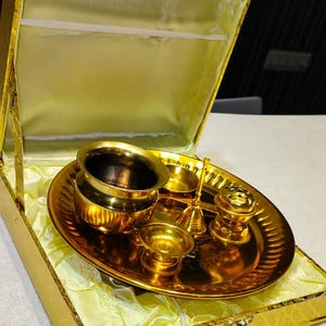 May include: A gold-toned metal puja thali set arranged on a circular tray, including a small bowl, a bell, and a diya, all housed within a matching gold-colored presentation box lined with pale yellow satin fabric for storage and display.