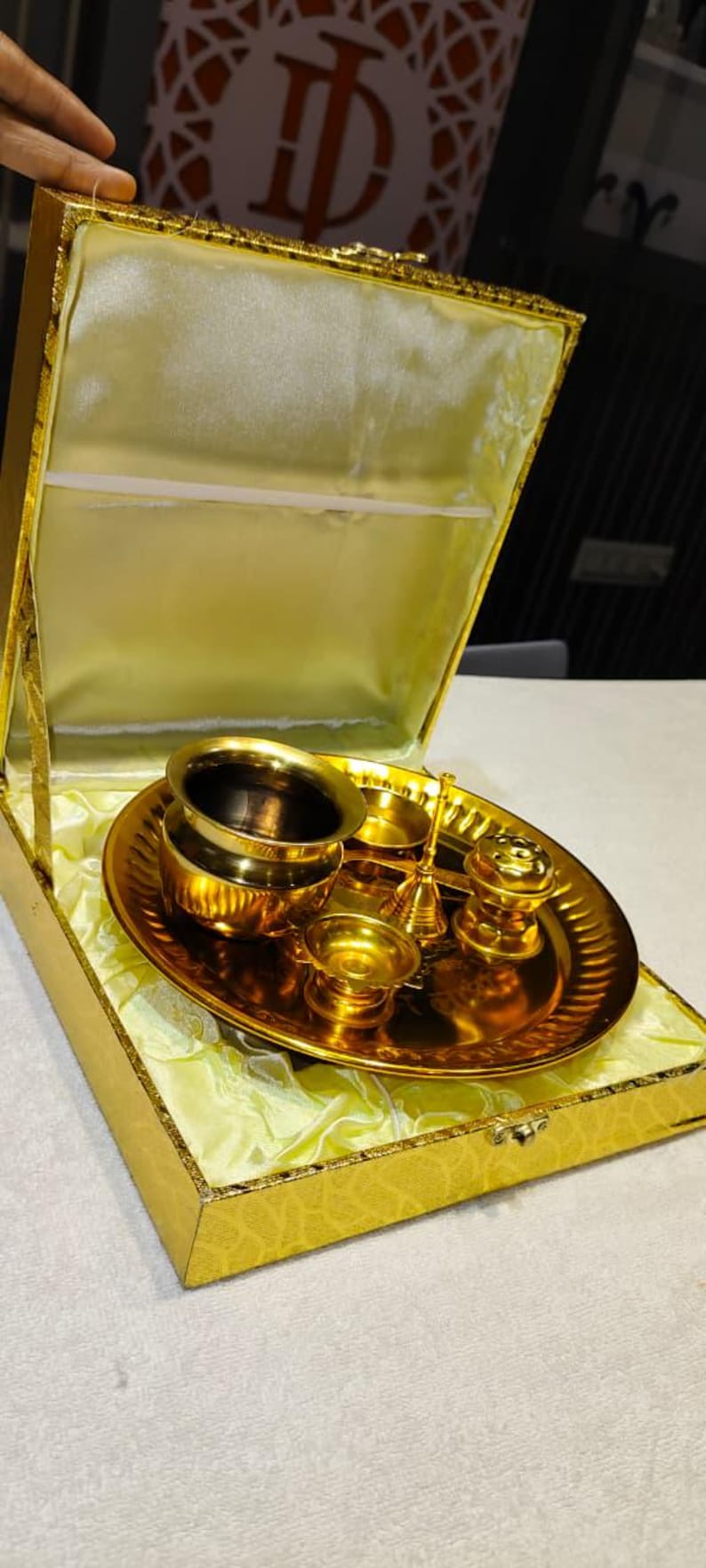May include: A gold-toned metal puja thali set arranged on a circular tray, including a small bowl, a bell, and a diya, all housed within a matching gold-colored presentation box lined with pale yellow satin fabric for storage and display.