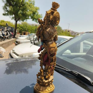 Puede incluir: Estatua dorada ornamentada de una deidad, posiblemente hind&uacute;. La figura est&aacute; adornada con tallas detalladas, una corona y prendas fluidas en rojo y azul. La estatua est&aacute; sobre una base de flor de loto.