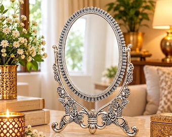 Puede incluir: Un espejo de mesa ovalado con un marco de tono plateado que presenta detalles florales y de hojas en relieve. El espejo se apoya en un soporte decorativo con pies curvos y motivos de rosas, permitiendo ajustar la inclinación del cristal.