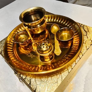 May include: A gold-toned metal puja thali set featuring a round tray with a scalloped edge, a small bowl, a larger cup, a spoon, a bell, and a small incense burner. The tray is engraved with script and rests on a matching gold-patterned box.