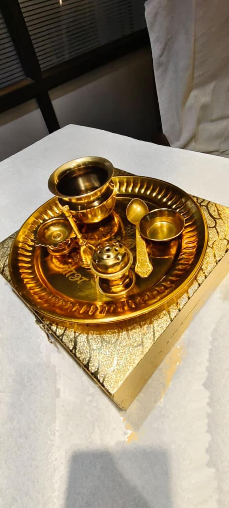 May include: A gold-toned metal puja thali set featuring a round tray with a scalloped edge, a small bowl, a larger cup, a spoon, a bell, and a small incense burner. The tray is engraved with script and rests on a matching gold-patterned box.