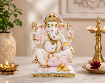 Puede incluir: Una estatua blanca de Ganesha con detalles en dorado y rosa, sentada sobre una base de flor de loto rosa. La deidad tiene cuatro brazos, sostiene un tridente y un aguijón, y presenta una pequeña figura de ratón en la base con adornos dorados.