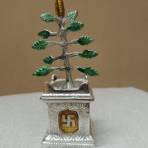 May include: A silver-toned metal decorative sculpture featuring a stylized tree with green painted leaves and a gold-colored top element. The tree is set into a square silver base with intricate embossed patterns and a central gold square with a swastika.